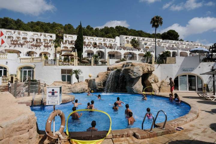 Un grupo de niños juega al waterpolo en una piscina con los apartamentos del resort al fondo.