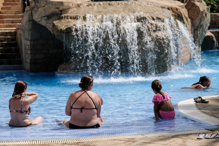Varias personas se sientan en la orilla de una piscina, cerca de una de las cascadas de la zona acuática.