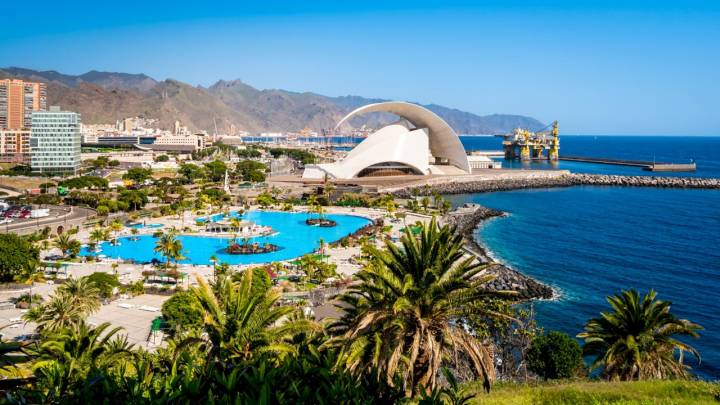 The iconic Auditorio de Tenerife auditorium gleaming in the sunlight and embodies the allure of this coastal capital city surrounded by the Anaga mountains, harbor and a tropical pool with palm trees.